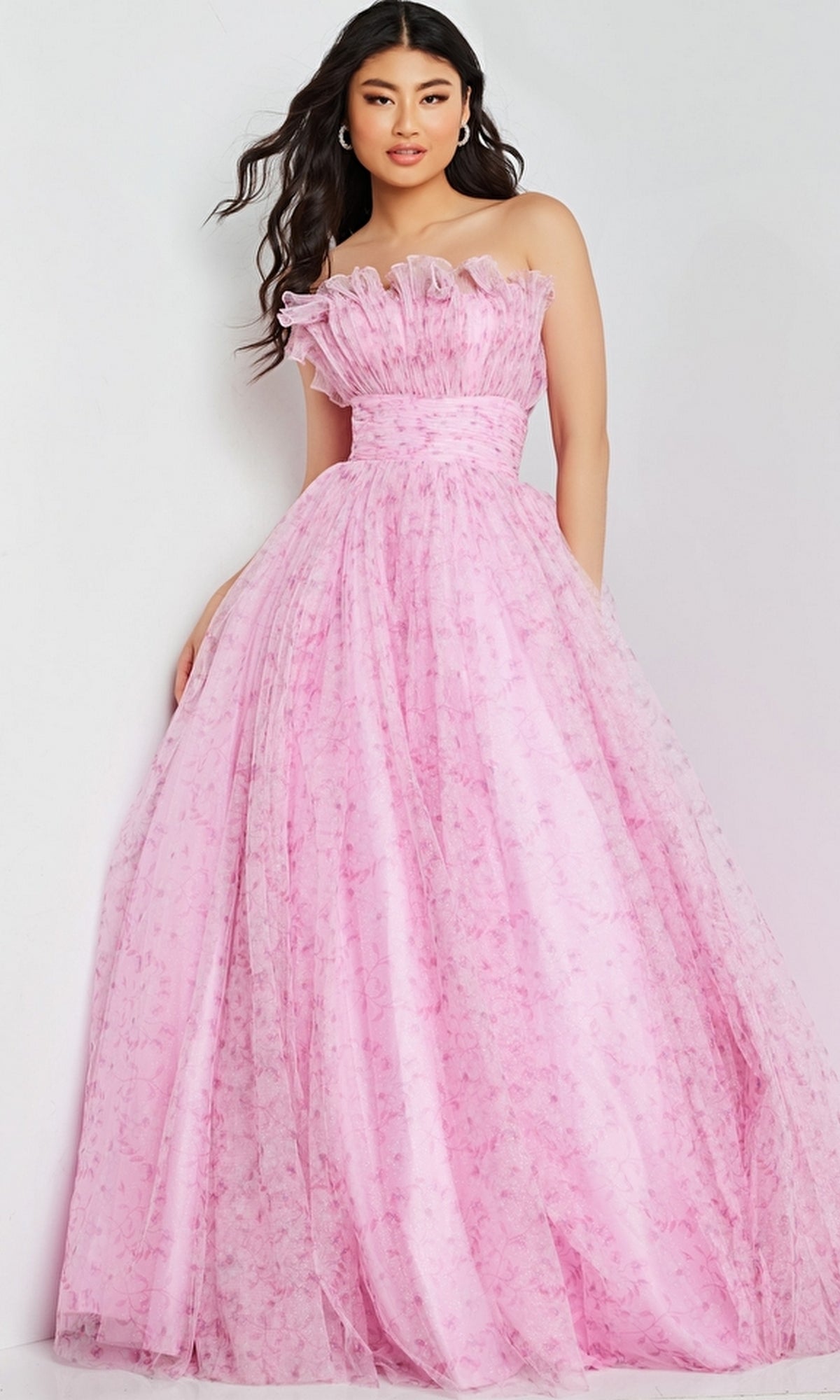 A woman wearing the Jovani 26209 long prom dress—a strapless, floor-length pink ball gown with ruffle neckline detail and floral patterns—stands against a light-colored background.